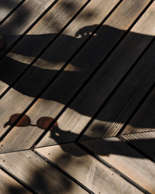 Shadow of person and sunglasses on wooden deck with angled planks creating a geometric pattern in light and dark tones.