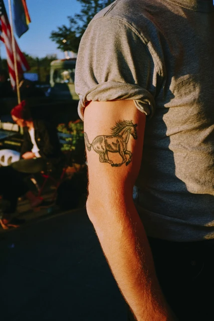 Close-up of a person's arm with a horse tattoo, wearing a grey t-shirt, set outdoors, showcasing the tattoo art in natural light.