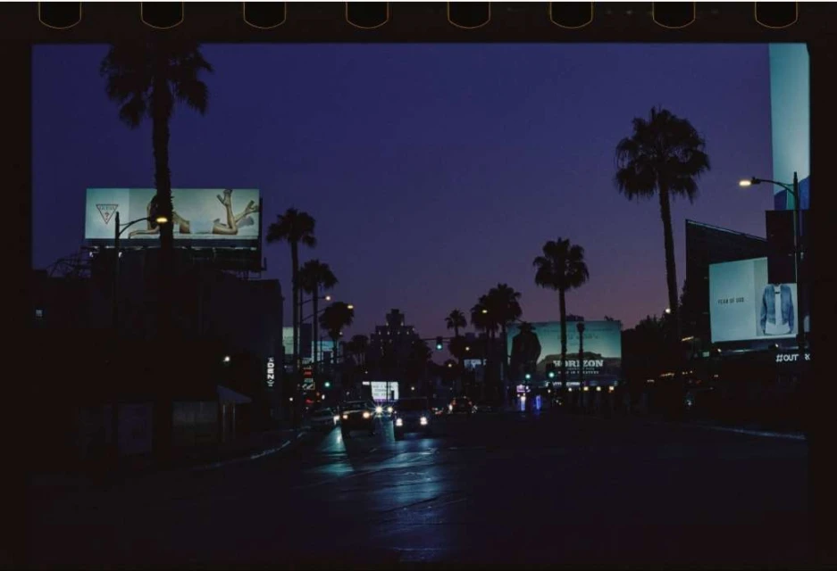 Urban twilight scene featuring palm trees, billboards, and streetlights lining a city street at dusk, creating a vibrant yet calming atmosphere.