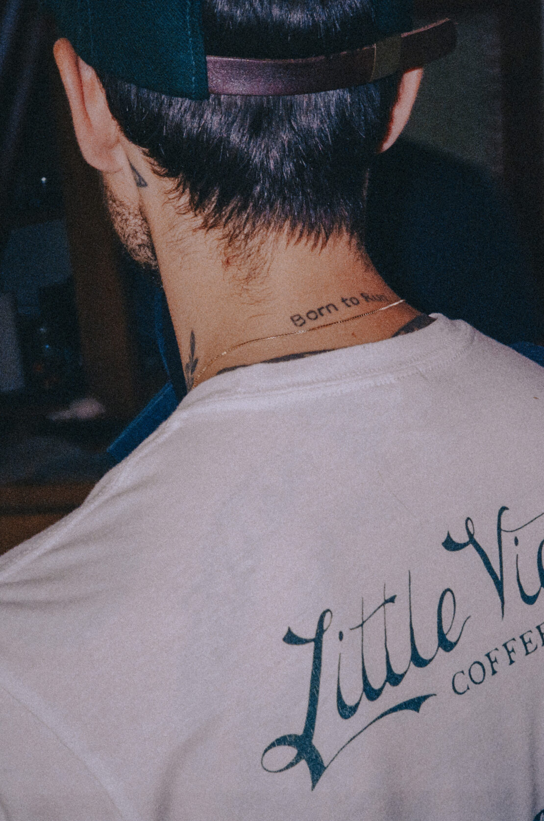 Adult male with dark hair and baseball cap, back to camera, wearing white t-shirt with Little Vices Coffee logo, neck tattoo visible.