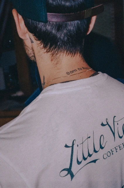 Adult male with dark hair and baseball cap, back to camera, wearing white t-shirt with Little Vices Coffee logo, neck tattoo visible.