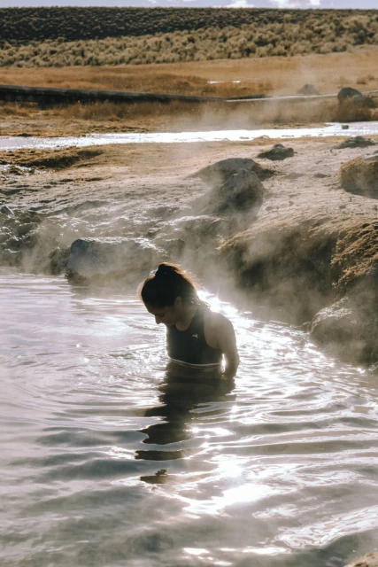 Woman in a black top soaks in a geothermic pool surrounded by desert landscape, under a warm, sunny sky in an outdoor setting.