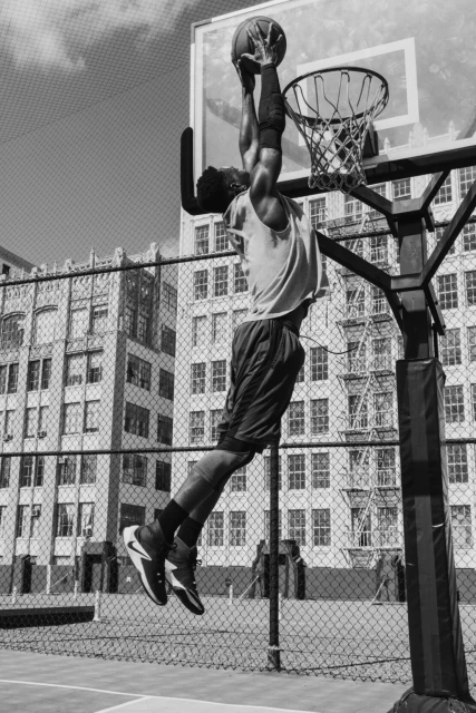 Athletic man in mid-air slam dunk on outdoor basketball court in black and white, featuring hoop, net, building and clear sky.