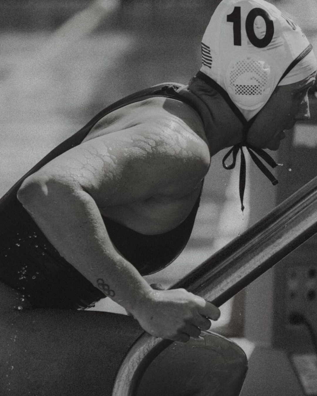 Black and white photo of a woman in a water polo cap with the number 10, gripping a wooden object, with an Olympic rings tattoo visible.