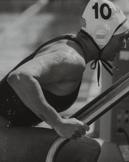 Black and white photo of a woman in a water polo cap with the number 10, gripping a wooden object, with an Olympic rings tattoo visible.