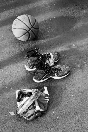 Monochrome overhead shot of a basketball, a pair of high-top sneakers, and folded athletic shorts on pavement.