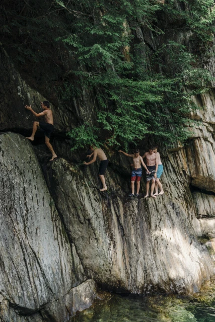 Group of young men climb and gather on a rocky cliff face with lush green trees above; swimming area below on a sunny day.
