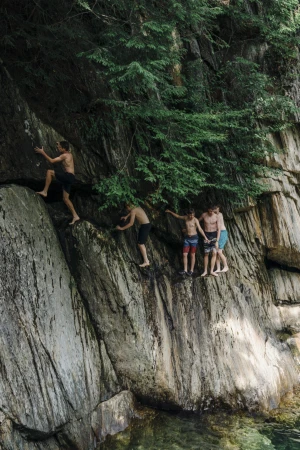 Group of young men climb and gather on a rocky cliff face with lush green trees above; swimming area below on a sunny day.