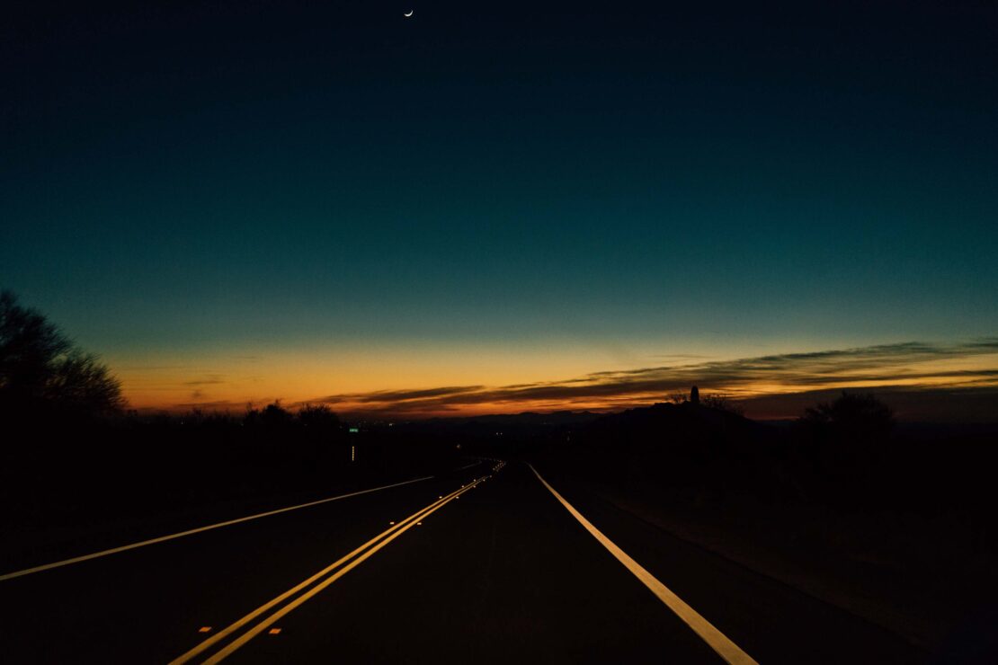 Dark asphalt road stretches into the distance under a twilight sky with an orange horizon and a crescent moon above. Trees in silhouette.