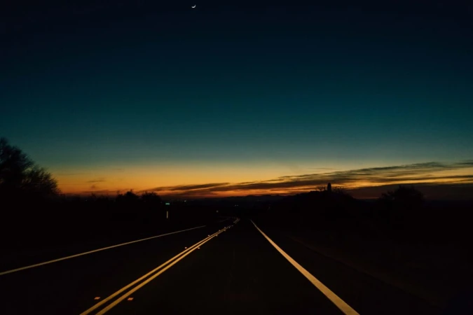 Dark asphalt road stretches into the distance under a twilight sky with an orange horizon and a crescent moon above. Trees in silhouette.