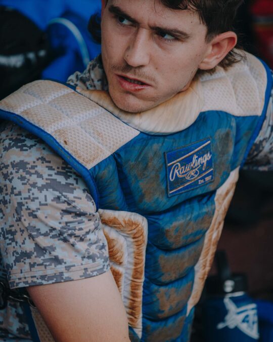 Close-up of baseball catcher wearing protective chest gear, posing indoors with serious expression, in medium shot at eye-level.