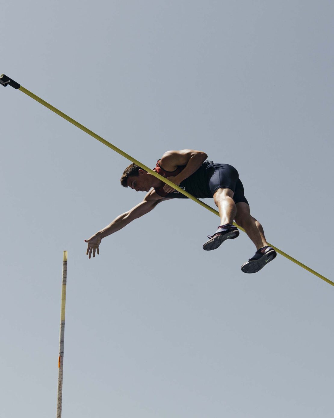 Male pole vaulter mid-air over the bar, athletic competition, blue sky background, intense concentration, muscular build, dynamic composition.
