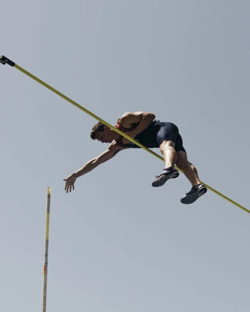 Male pole vaulter mid-air over the bar, athletic competition, blue sky background, intense concentration, muscular build, dynamic composition.