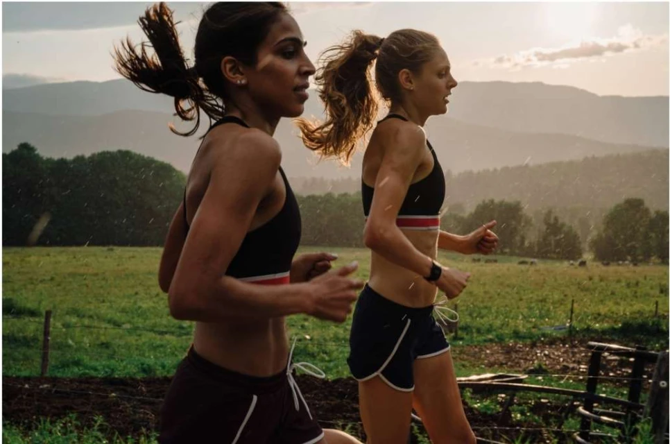 Two athletic women are running outdoors in sports bras and shorts, showcasing fitness and health against a blurred background of mountains.