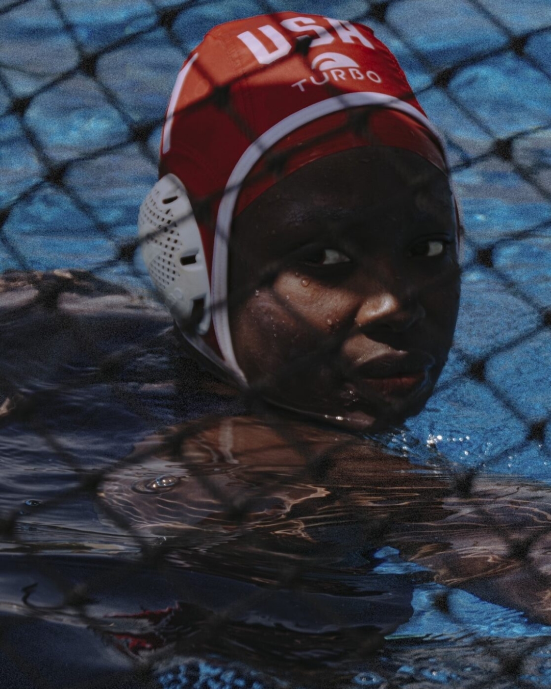 Close-up of a dark-skinned woman wearing a red water polo cap with the text 'USA Turbo' in a pool, with a net visible in the foreground.