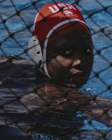 Close-up of a dark-skinned woman wearing a red water polo cap with the text 'USA Turbo' in a pool, with a net visible in the foreground.