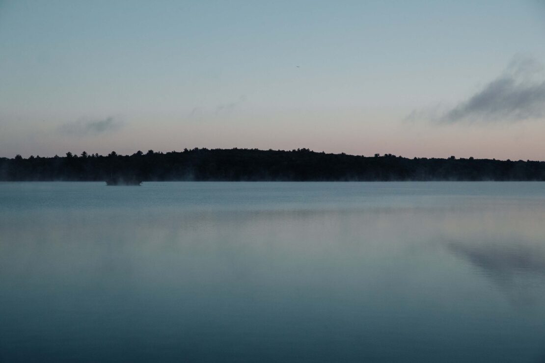 Calm lake at dusk with silhouette of trees and a small island, reflections creating a mirror-like surface under a gradient sky.