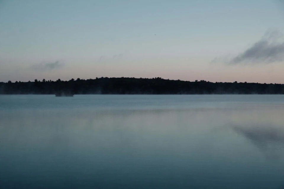 Calm lake at dusk with silhouette of trees and a small island, reflections creating a mirror-like surface under a gradient sky.