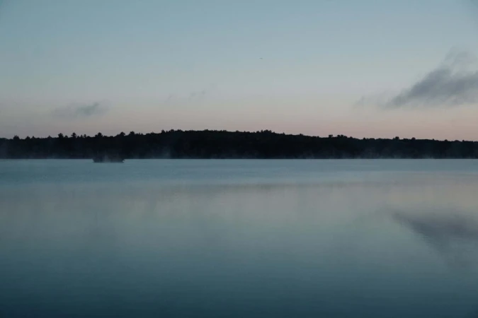 Calm lake at dusk with silhouette of trees and a small island, reflections creating a mirror-like surface under a gradient sky.
