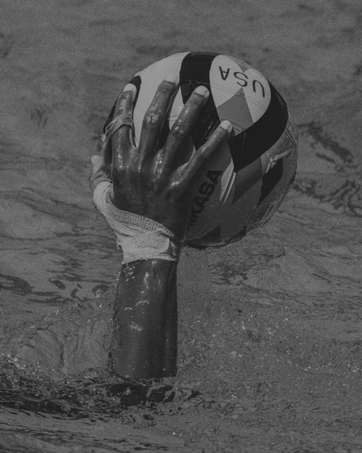 Monochrome shot of a hand holding a USA water polo ball above rippling water with a Mikasa logo visible on the ball's surface.