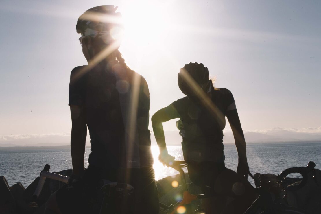 Two cyclists in helmets silhouetted by bright sun near ocean. One stands with bike, other sits on theirs. Sky and horizon visible.
