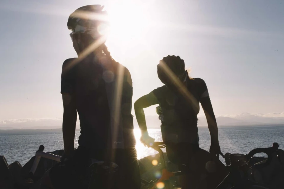 Two cyclists in helmets silhouetted by bright sun near ocean. One stands with bike, other sits on theirs. Sky and horizon visible.