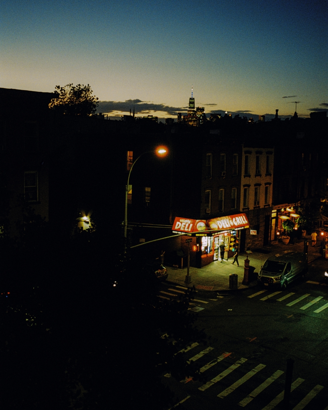 Night cityscape featuring a deli on a corner, illuminated by streetlights, with a view of city buildings under the evening sky.