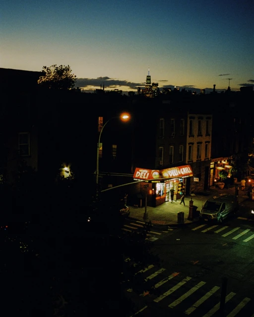 Night cityscape featuring a deli on a corner, illuminated by streetlights, with a view of city buildings under the evening sky.