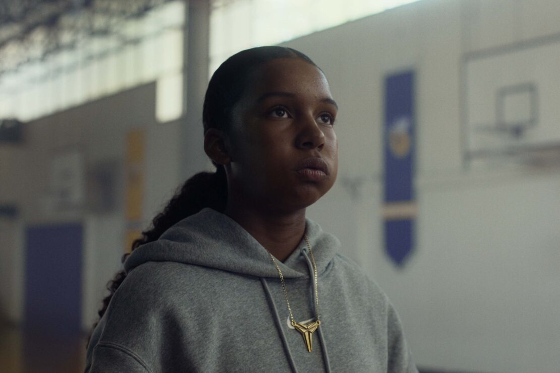 Young woman in a grey hoodie wearing a gold necklace inside a gymnasium by photographer Emily Maye