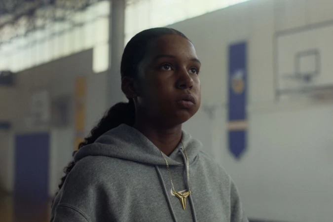 Young woman in a grey hoodie wearing a gold necklace inside a gymnasium by photographer Emily Maye