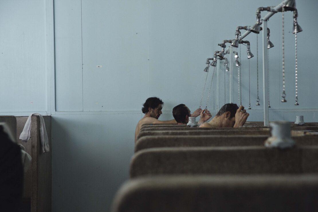 Several adult males stand in an indoor shower room with metal fixtures and aged light blue walls, engaging in bathing.