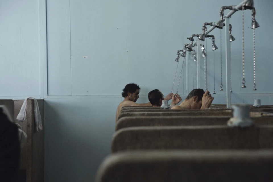 Several adult males stand in an indoor shower room with metal fixtures and aged light blue walls, engaging in bathing.