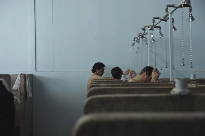 Several adult males stand in an indoor shower room with metal fixtures and aged light blue walls, engaging in bathing.
