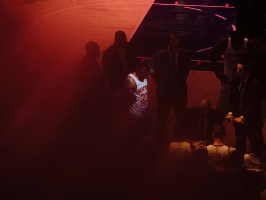 A basketball player in uniform stands on the court, surrounded by coaches and teammates in a reddish indoor basketball stadium.