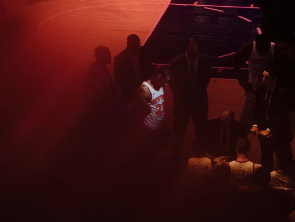 A basketball player in uniform stands on the court, surrounded by coaches and teammates in a reddish indoor basketball stadium.