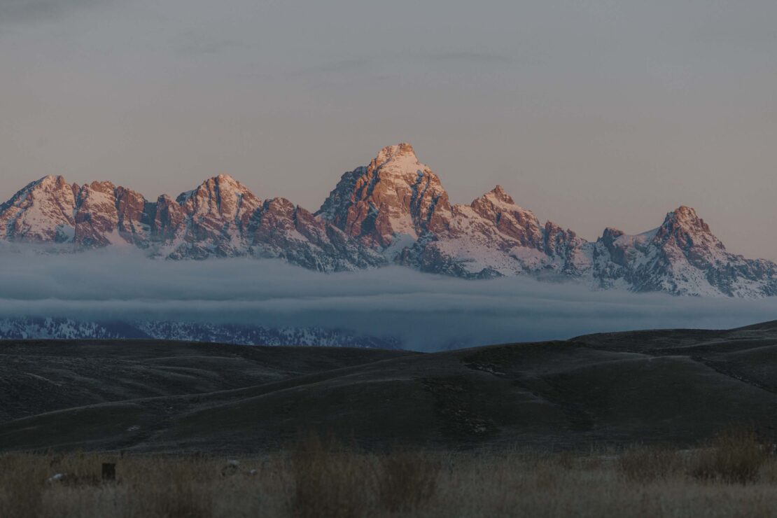 Scenic view of snow-capped mountain range partially obscured by clouds in a rural landscape, bathed in soft, early morning light.