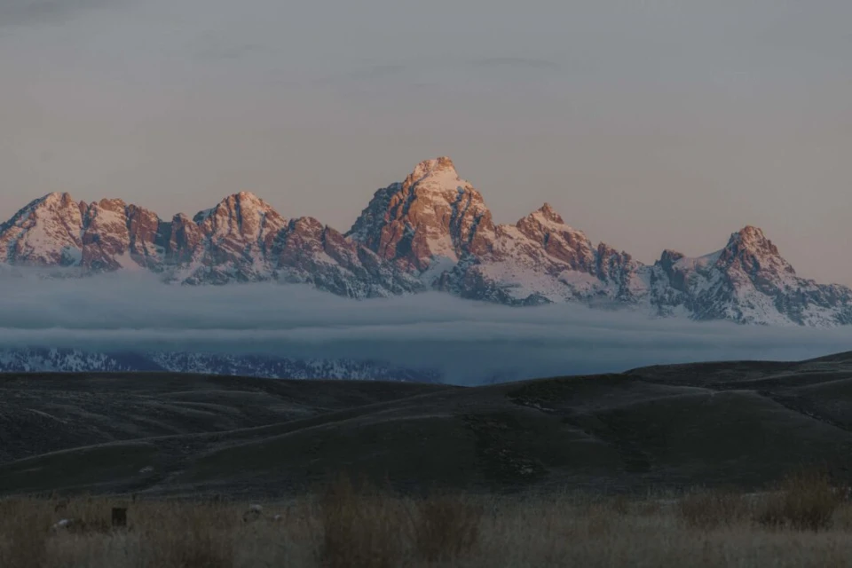 Scenic view of snow-capped mountain range partially obscured by clouds in a rural landscape, bathed in soft, early morning light.