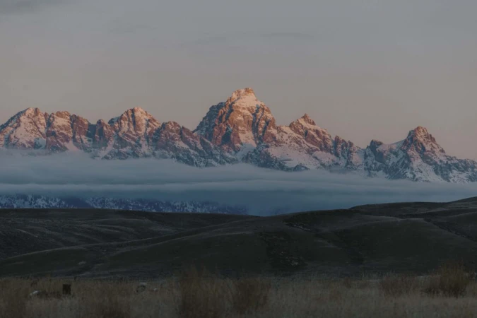 Scenic view of snow-capped mountain range partially obscured by clouds in a rural landscape, bathed in soft, early morning light.