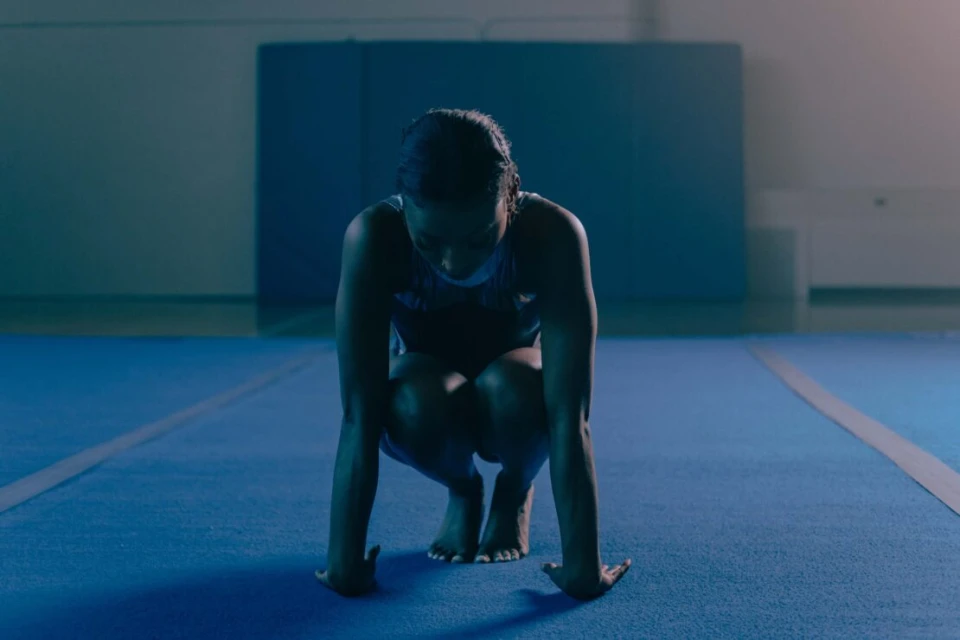 A gymnast is crouched on a blue mat in an indoor gymnasium, preparing for a routine with her hands pressed against the floor.