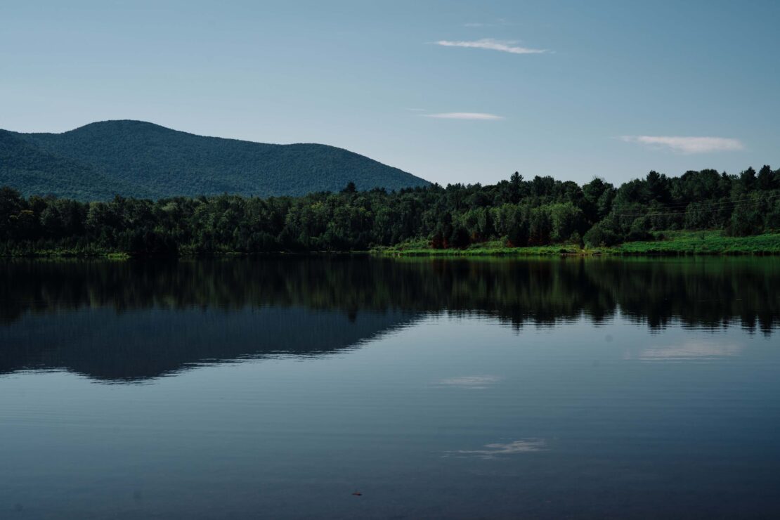 Calm dark blue lake reflecting a dense dark green forest and distant rolling mountain under a clear blue sky with thin clouds.