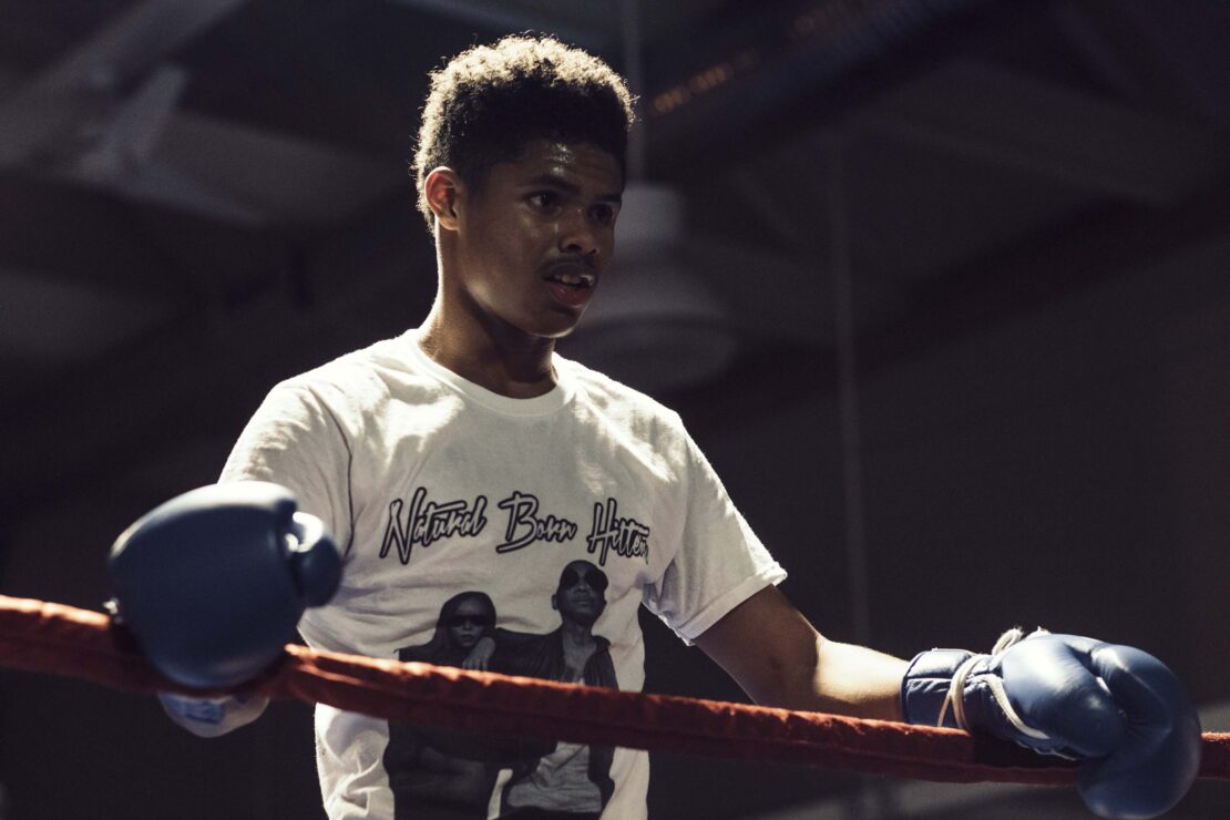 Young man with boxing gloves leans on ropes in a boxing ring, looking determined and focused during a training session.