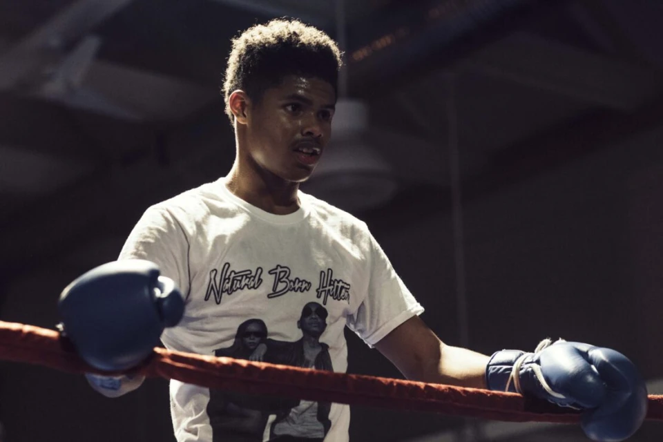 Young man with boxing gloves leans on ropes in a boxing ring, looking determined and focused during a training session.