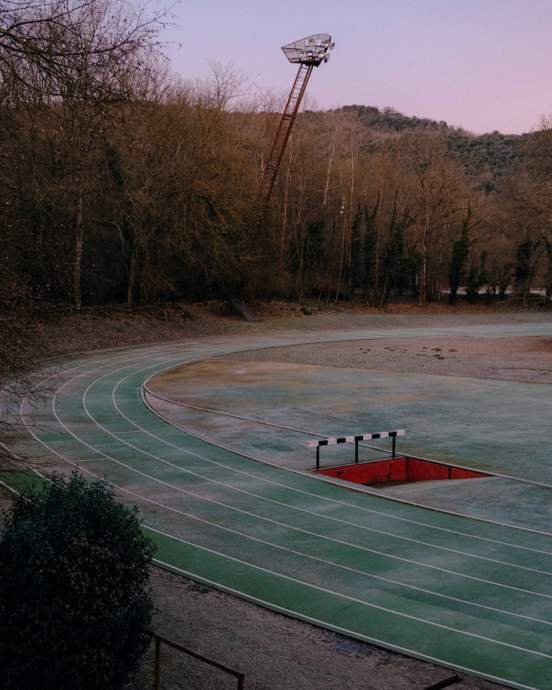 Running track with lanes, a hurdle, and a tall light stand, set against a backdrop of trees and a dusky pink sky.