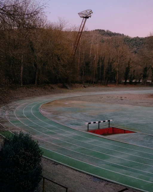 Running track with lanes, a hurdle, and a tall light stand, set against a backdrop of trees and a dusky pink sky.
