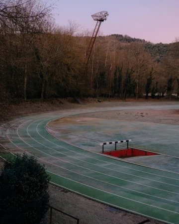 Running track with lanes, a hurdle, and a tall light stand, set against a backdrop of trees and a dusky pink sky.