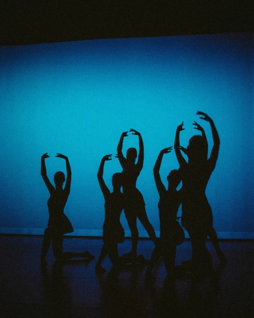 Silhouette of five women performing a dance routine in front of a light blue background on a dark stage. Low-key lighting with dark floor.