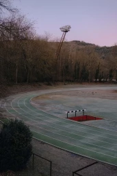 An empty green running track curves past a water jump pit under a pale dusk sky and sparse winter trees.
