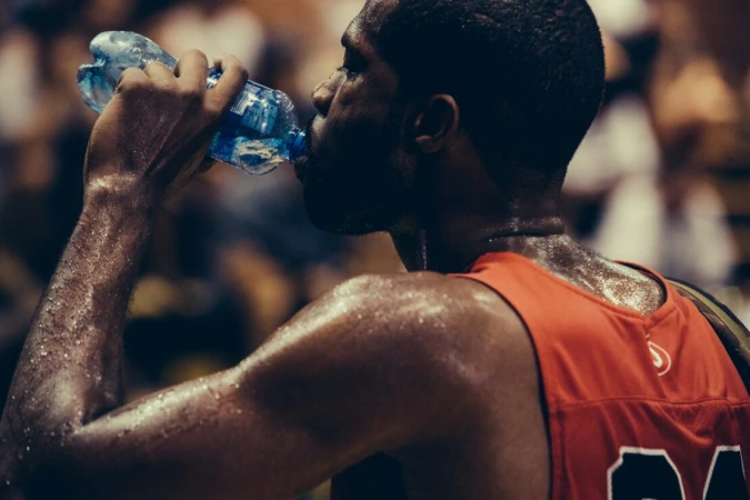 Sweaty athlete drinking water from a plastic bottle, wearing a red sports jersey in a dim interior setting.