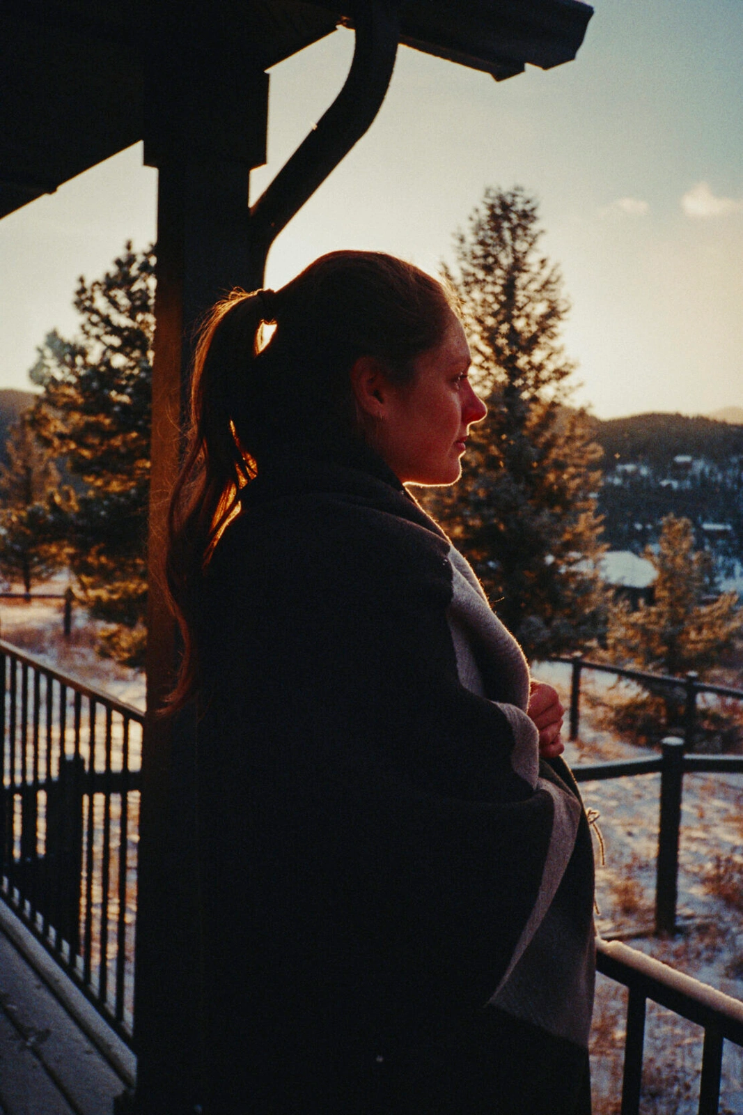 Profile of a woman wrapped in a dark blanket standing on a wooden deck at sunset with snowy pine trees in the background
