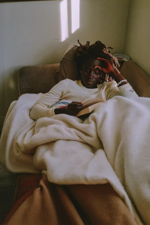 Black woman with dreadlocks relaxing on a sofa under a cream blanket reading a book